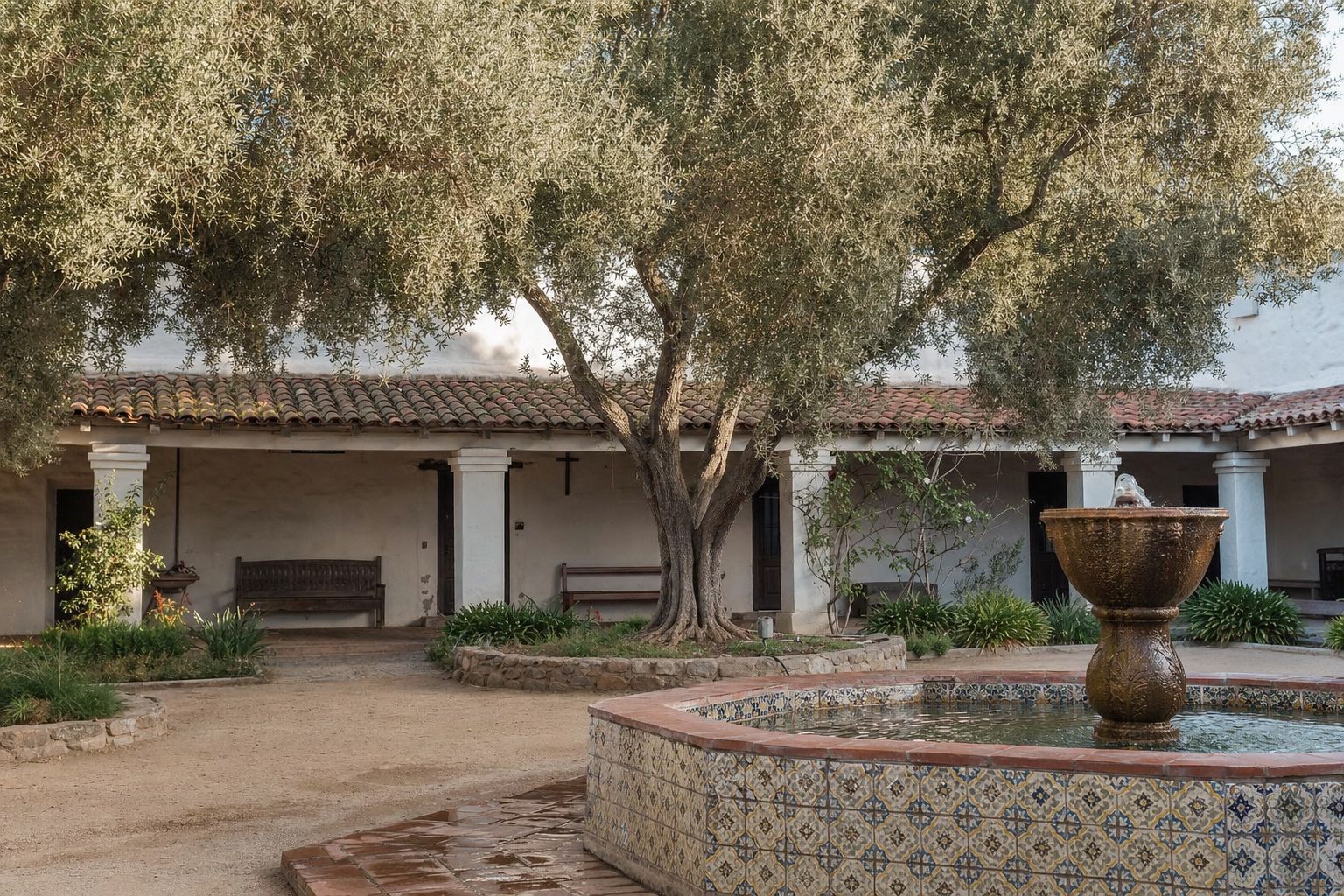 A whitewashed Spanish mission courtyard with a tiled fountain and mature olive trees